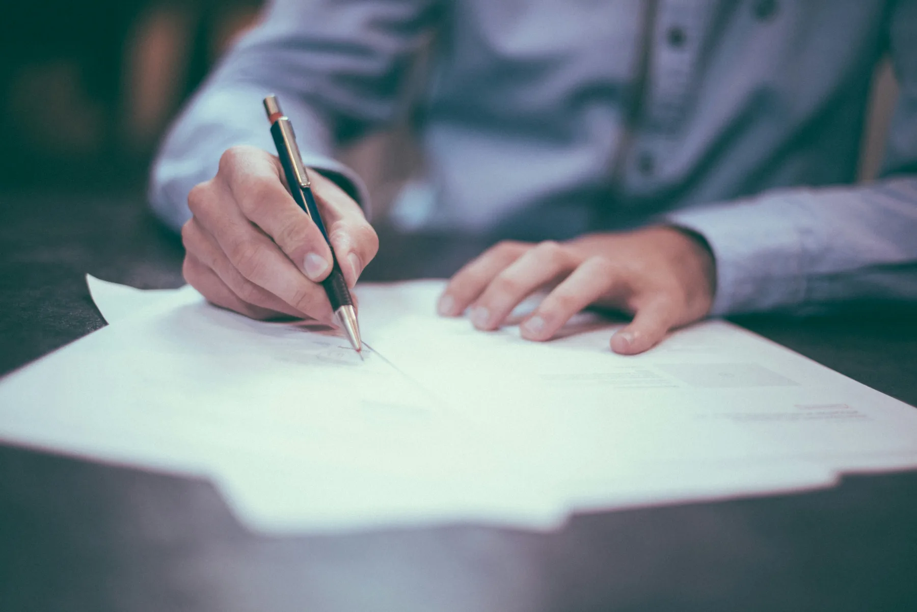 A person in a light blue shirt filling out and signing a paper form at a dark desk