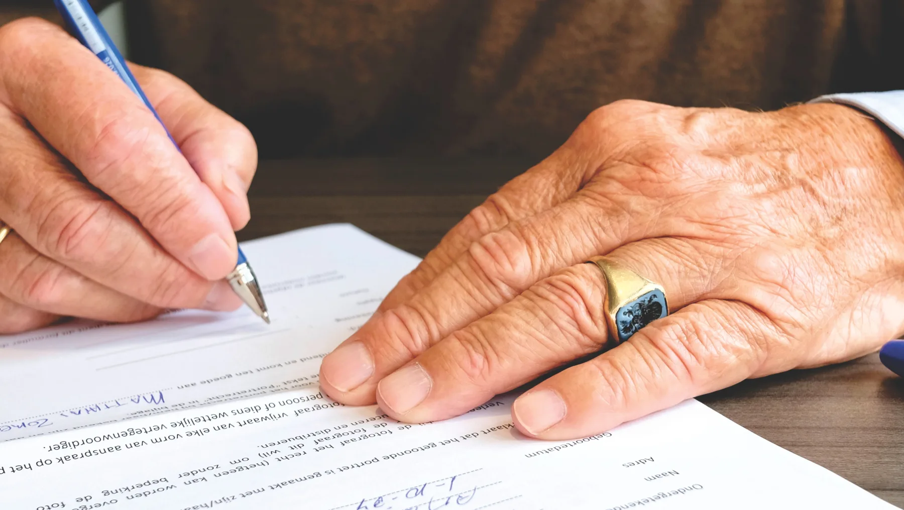 Close-up of an older man's hand signing a form with a pen while another hand holds the pages steady on a desk