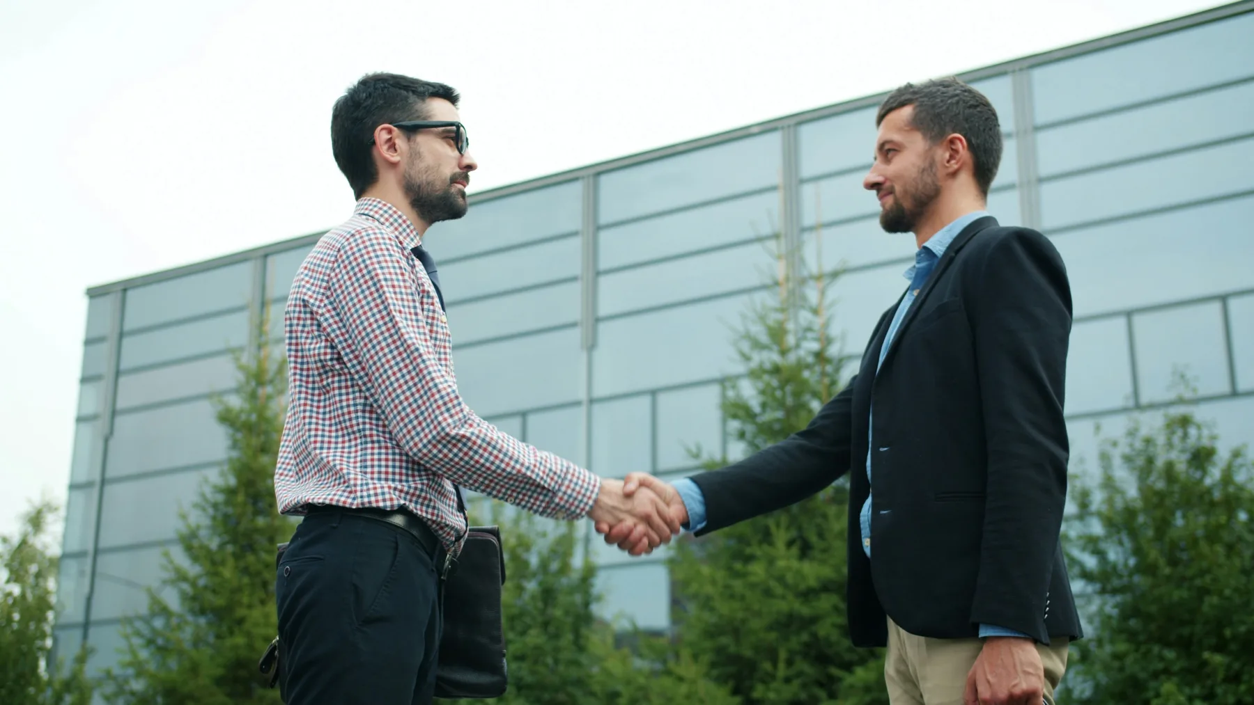 Two men in business attire shaking hands outdoors in front of a glass office building