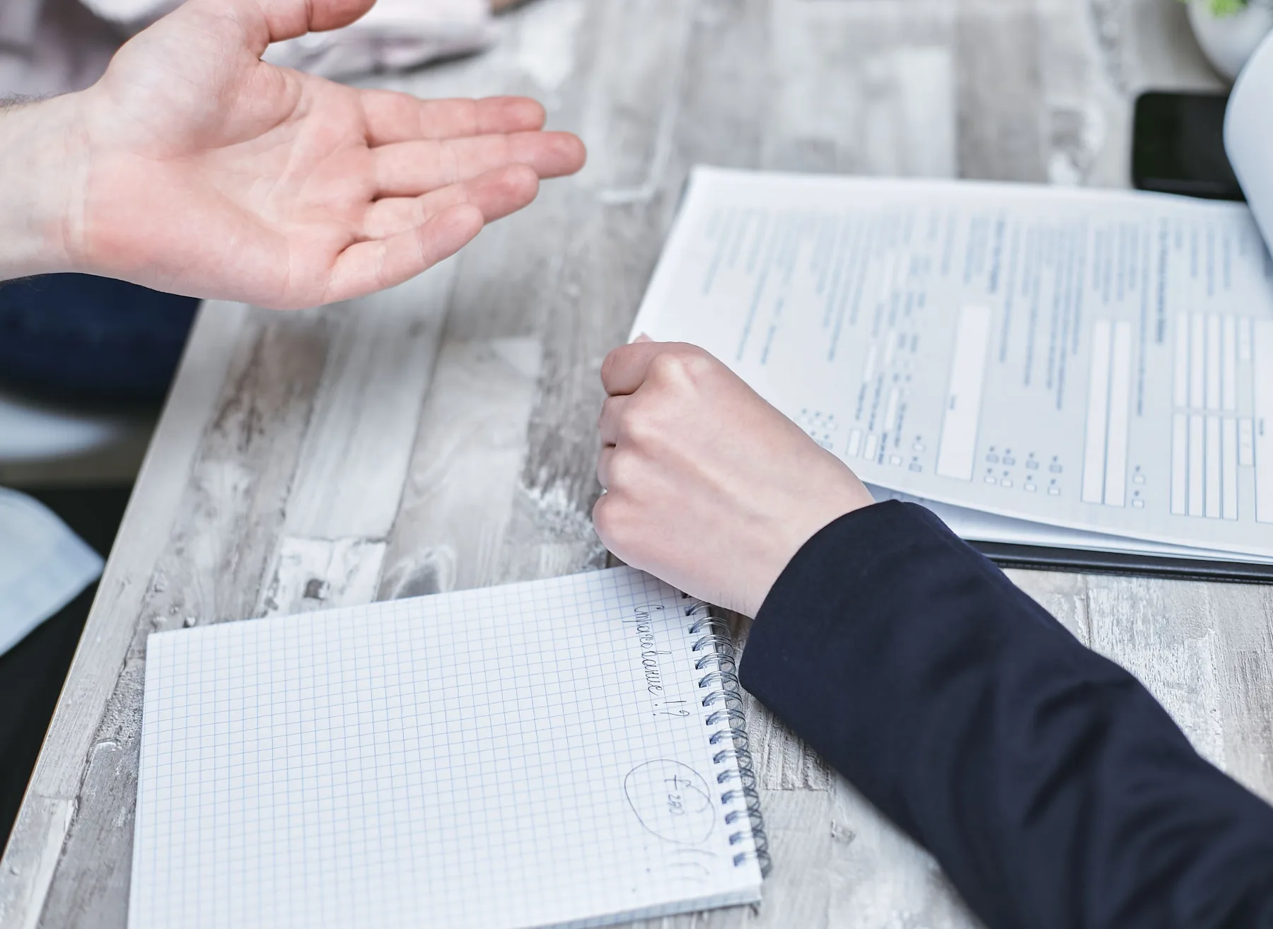 A person gesturing with an open palm while explaining a printed form, with a spiral notebook and pen nearby on a wooden table
