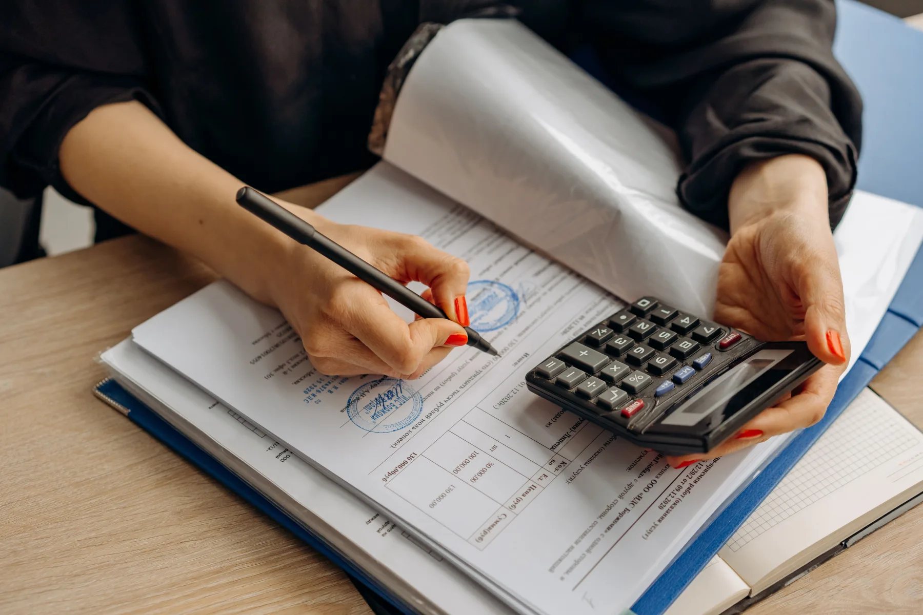 Woman using a calculator while reviewing a stamped official document with columns of figures, pen in hand