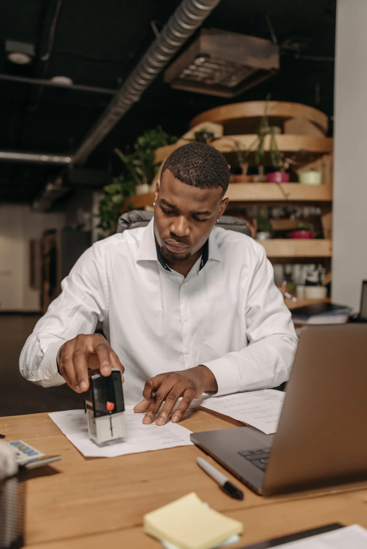 Man in a white shirt using a hand-held stamp to mark a document at his desk, laptop open beside him
