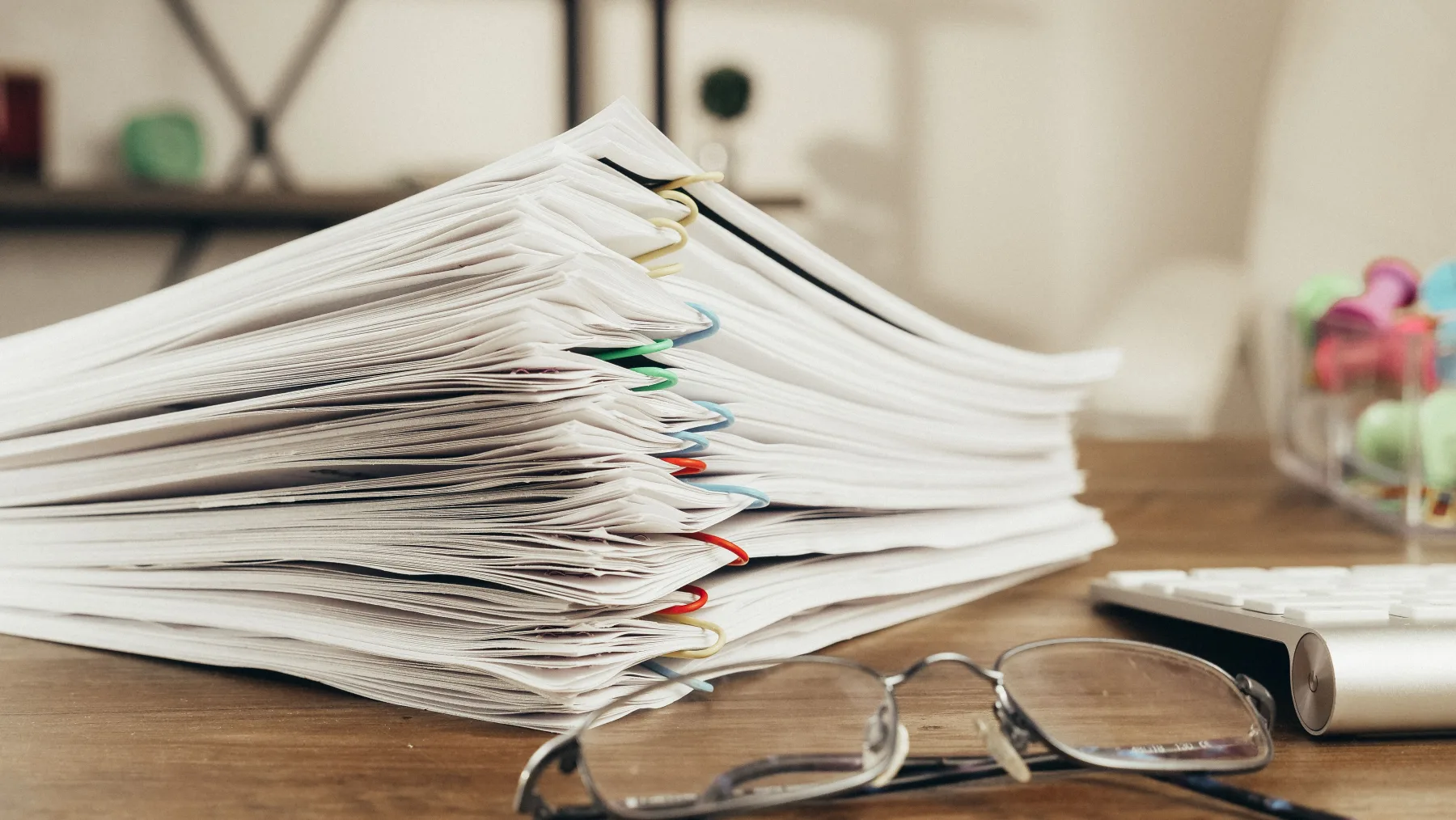 A thick stack of paper documents bound with colorful paper clips, reading glasses resting on the desk beside a keyboard
