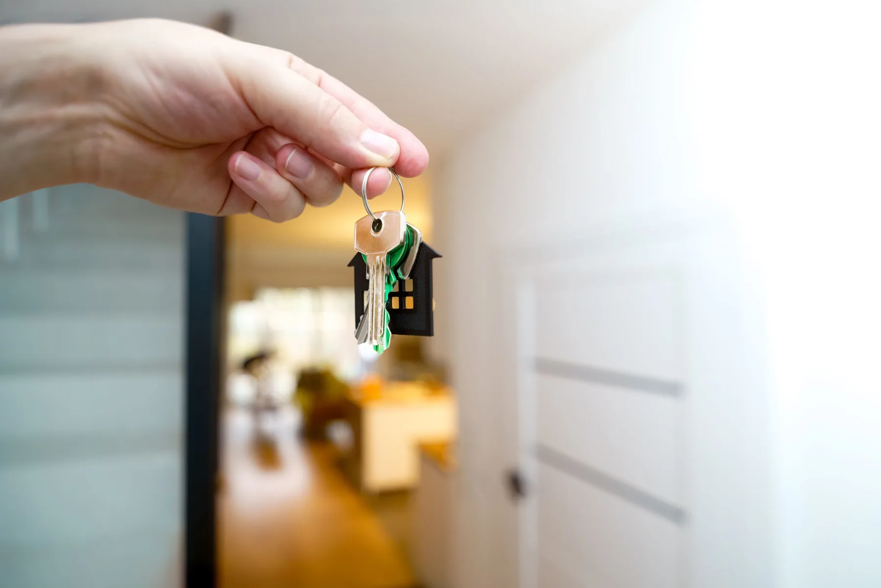 Hand holding a house-shaped keychain with keys in the doorway of a home, looking into a bright living area