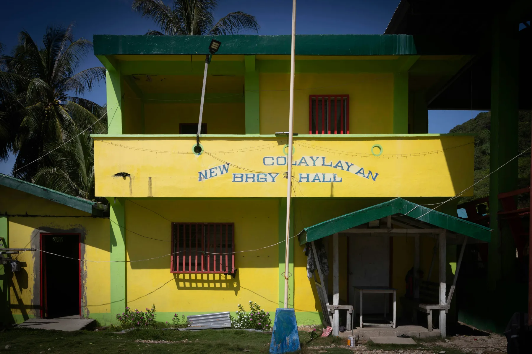 Bright yellow two-story barangay hall with a green roof and palm trees, signage reading 'New Colaylayan Brgy Hall'