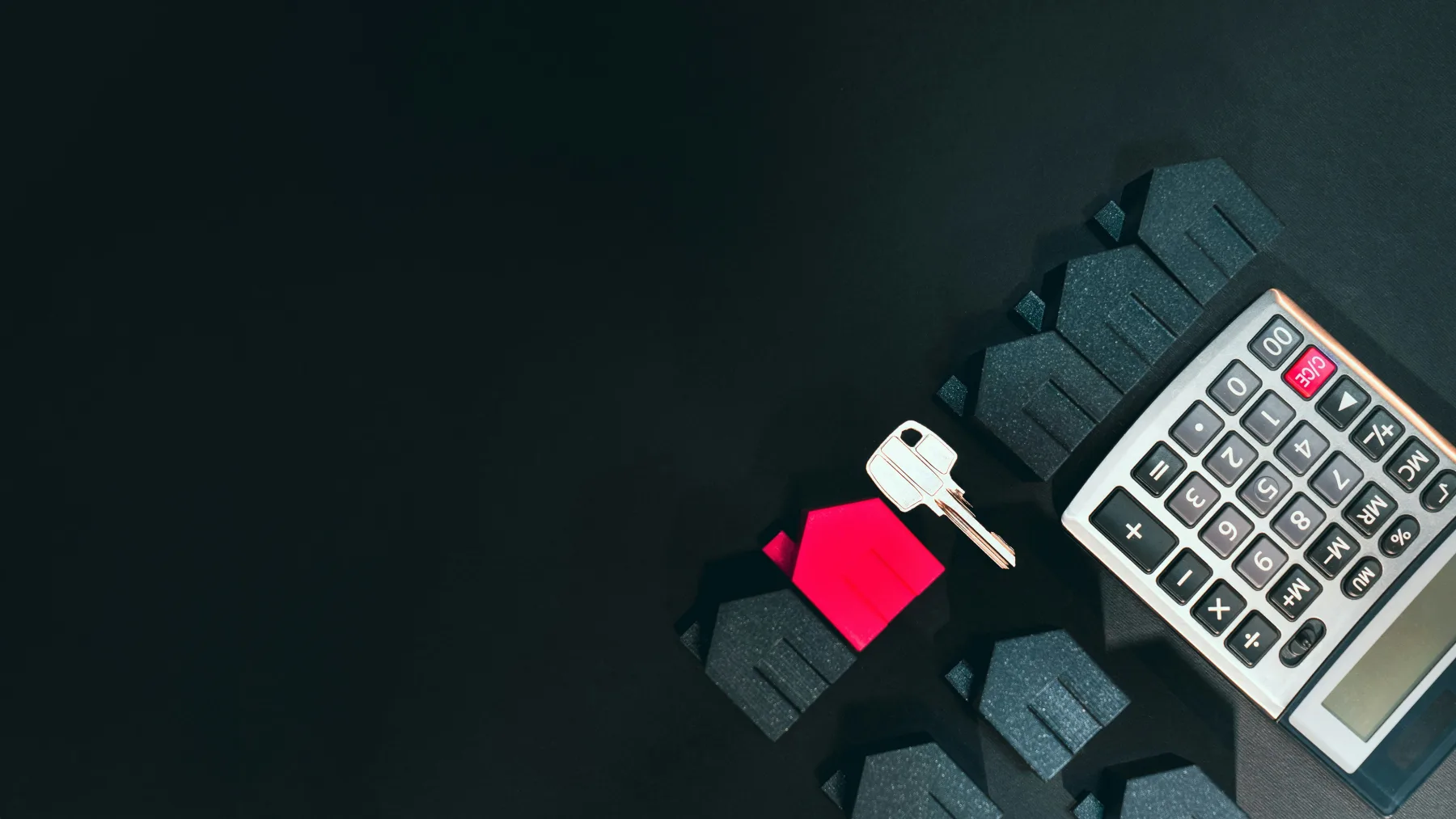 A small red house-shaped keychain with its key resting beside a calculator on a dark surface, with rows of dark toy houses nearby.