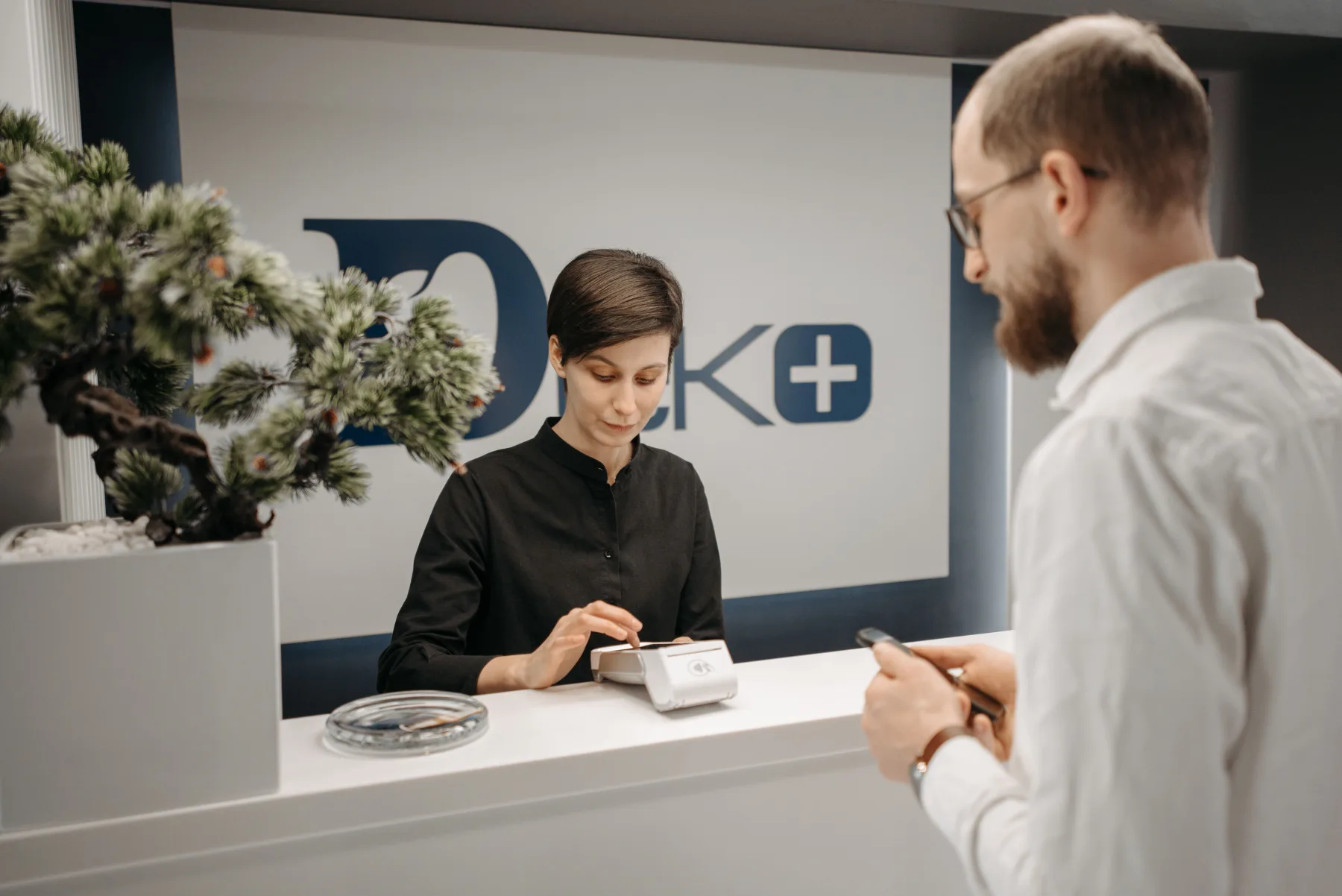 A staff member behind a reception counter processes a payment on a card terminal while a customer holds up his phone and card at a front desk.
