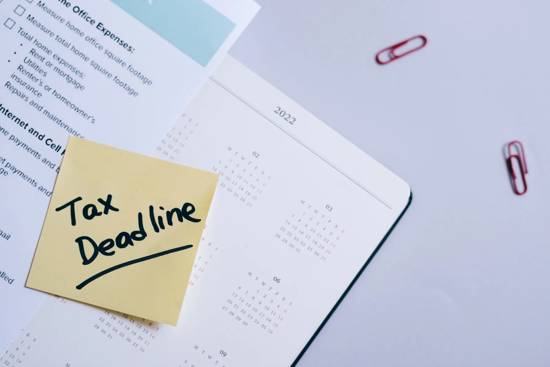 A yellow sticky note reading 'Tax Deadline' stuck on a page of a desk calendar, next to a home-expenses checklist and red paperclips.
