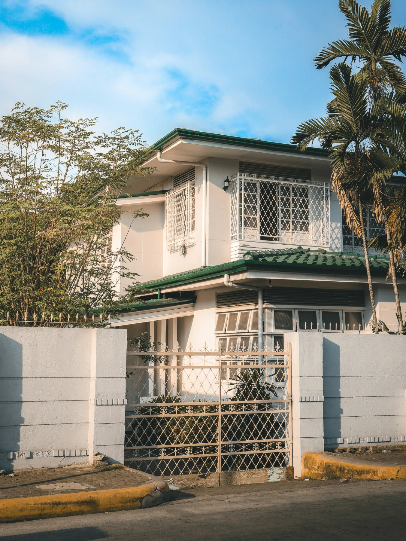 A two-story Philippine home with a green roof, iron window grilles, and a gated concrete perimeter wall behind palm trees.