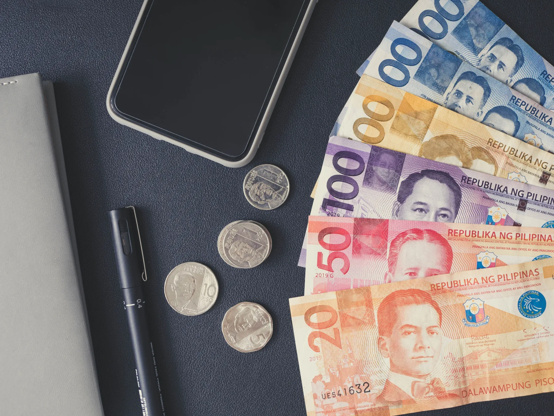 Philippine peso bills of several denominations fanned out next to loose coins, a notebook, a pen, and a smartphone on a dark table.