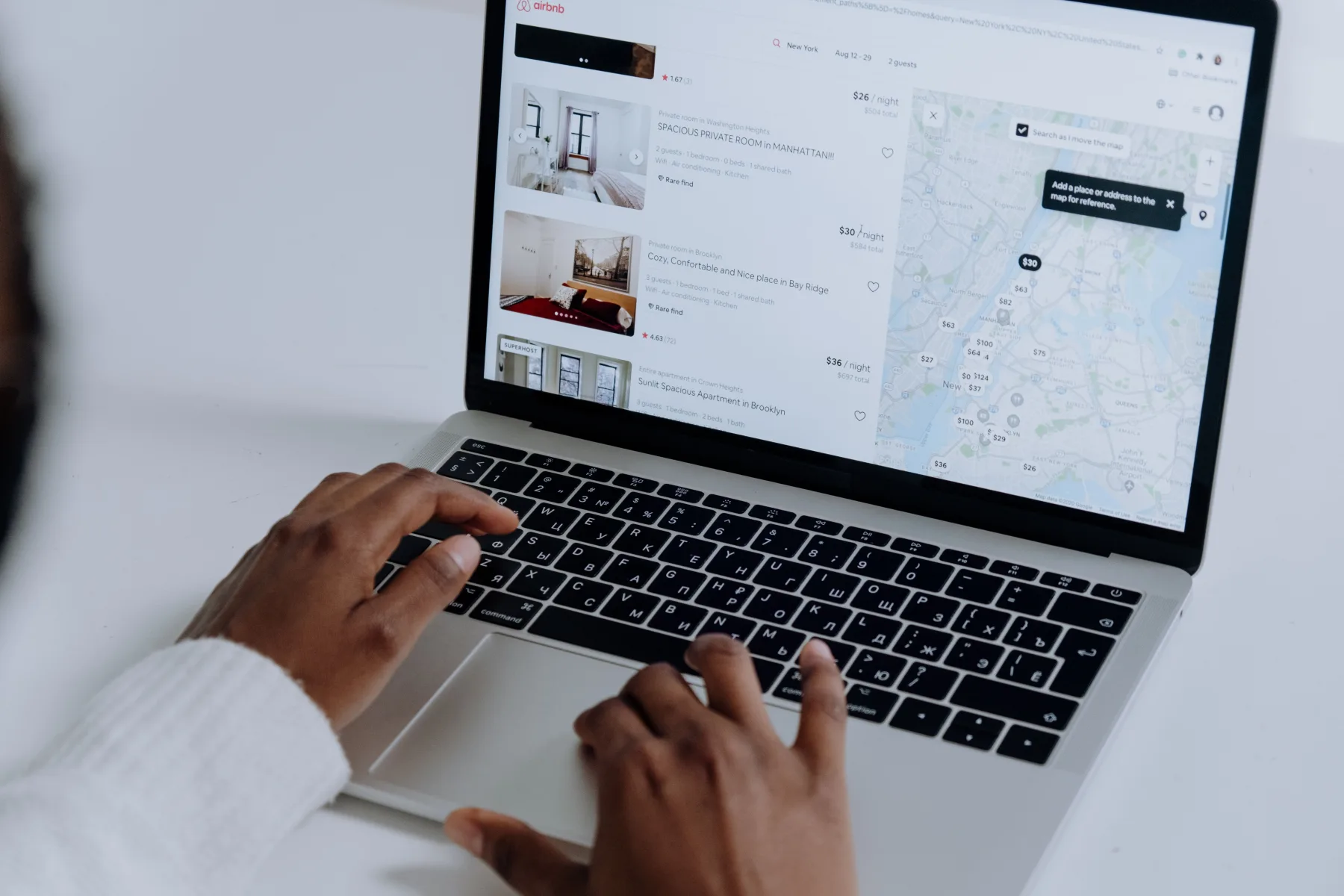 A laptop screen showing an Airbnb-style property search with listing photos and prices on the left and an interactive map with price pins scattered across a city on the right, with a person's hands on the keyboard and trackpad