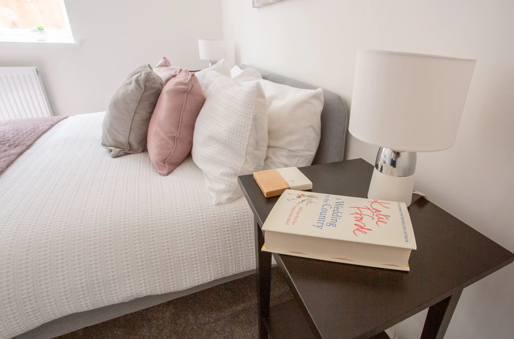 A cozy bedroom nightstand with a table lamp, two paperback books, and a marble coaster, set against pastel pink and grey pillows on a neatly made bed