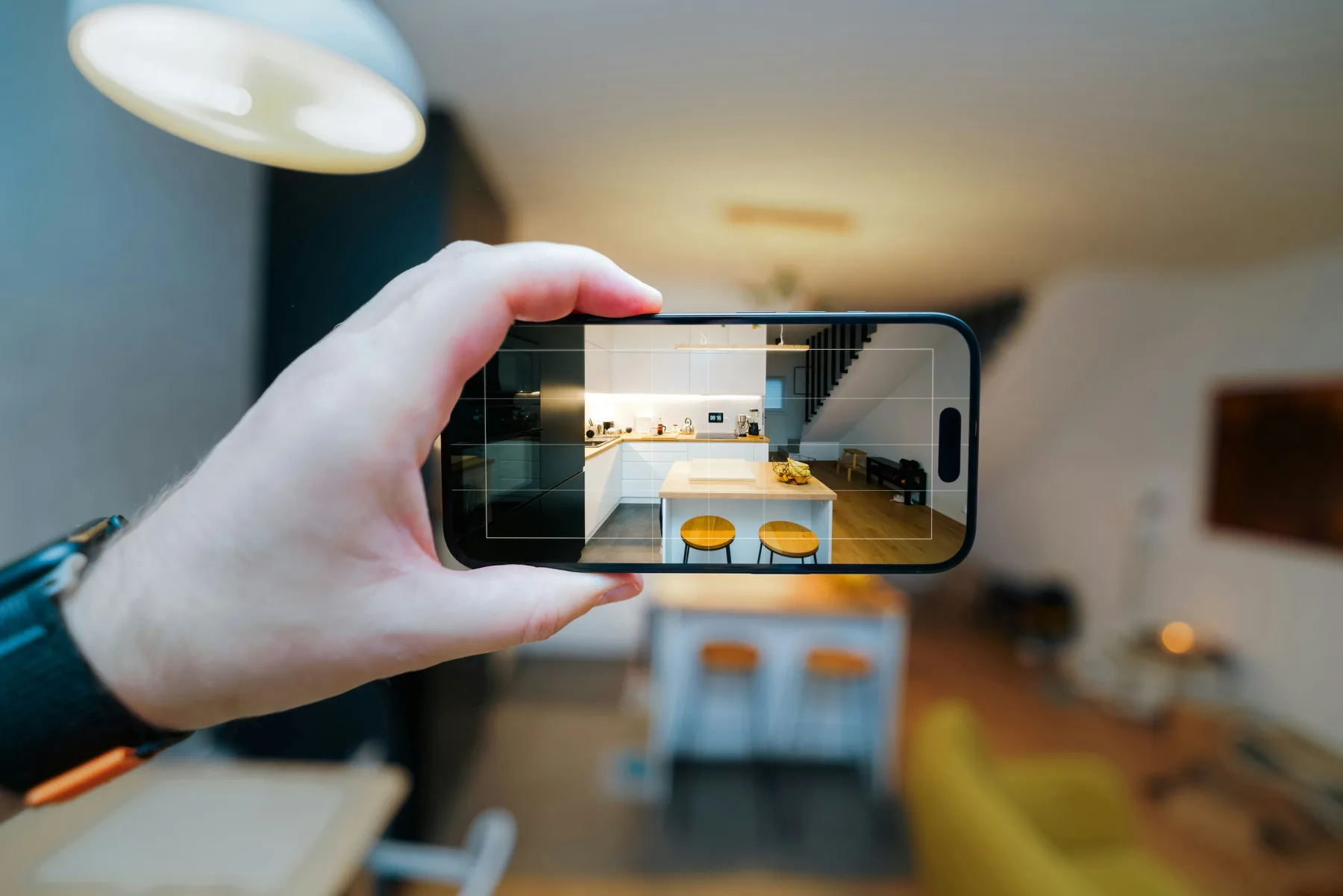 A hand holding up a smartphone with a camera grid overlay, framing a shot of a bright kitchen and dining area with wooden stools and a staircase in the background