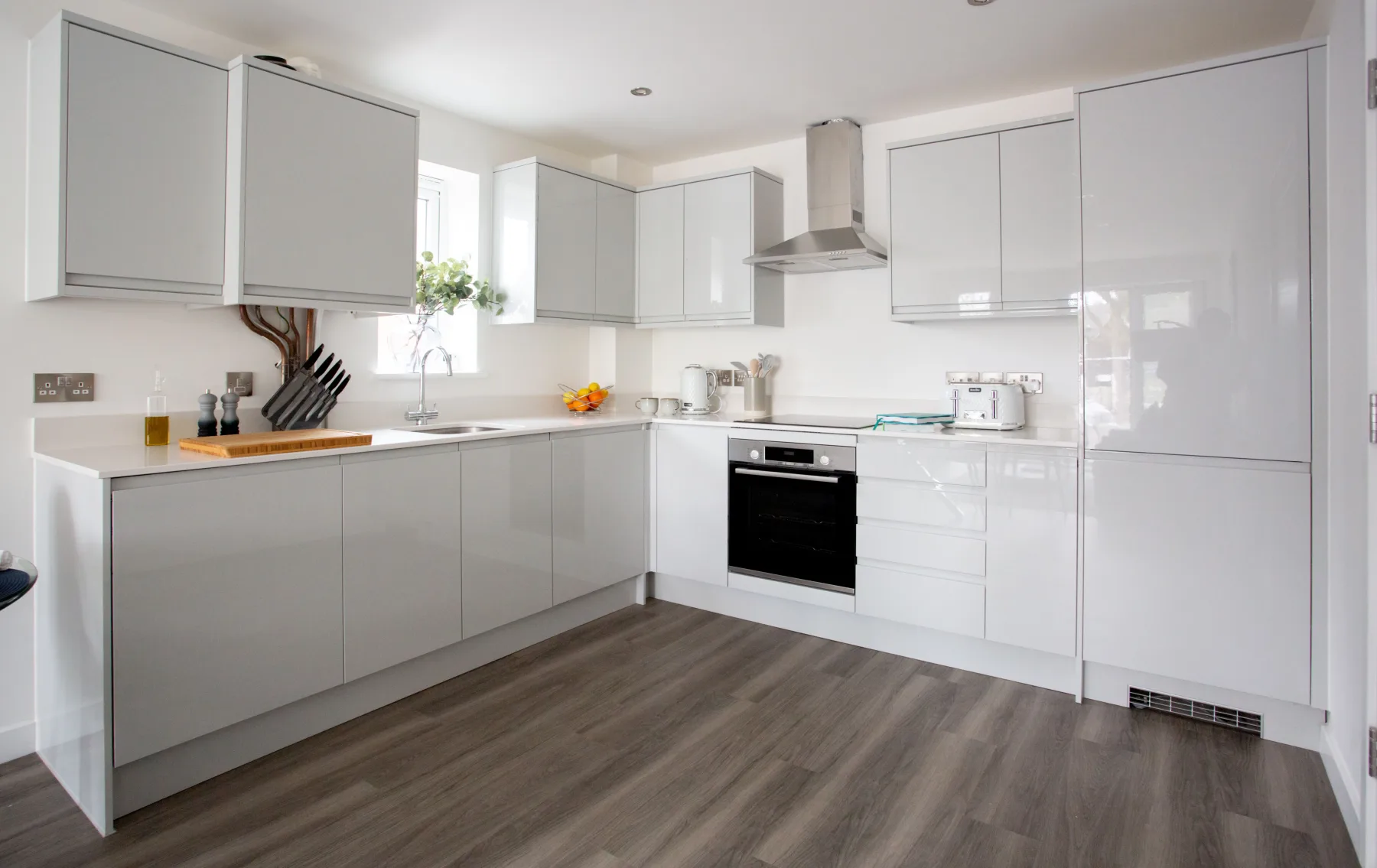 A bright, modern L-shaped kitchen with glossy light-grey cabinetry, a built-in oven, stainless range hood, and dark wood-look flooring