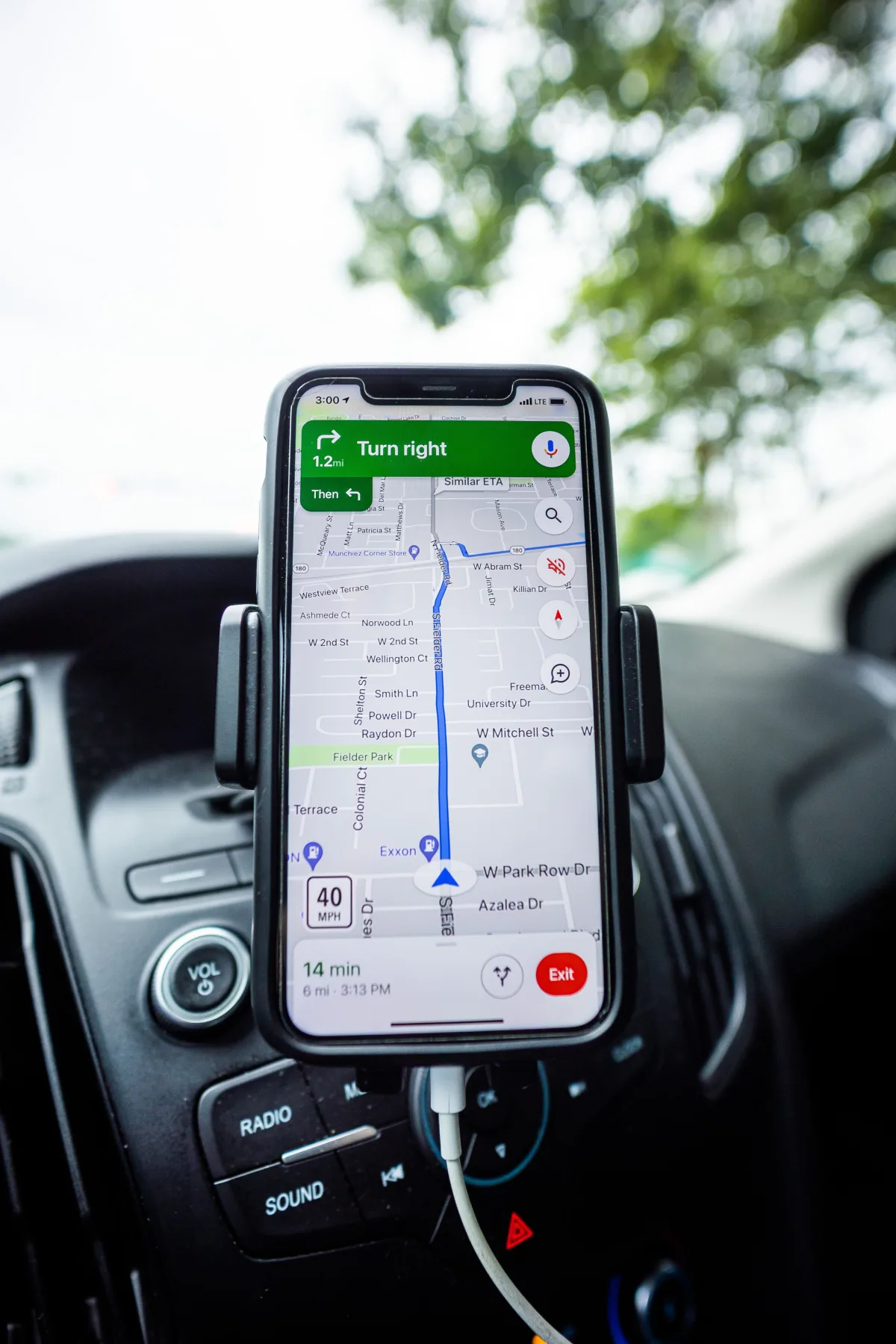 A smartphone mounted on a car dashboard showing turn-by-turn GPS navigation, a 14-minute ETA, and a 40 mph speed limit, with a tree visible through the windshield