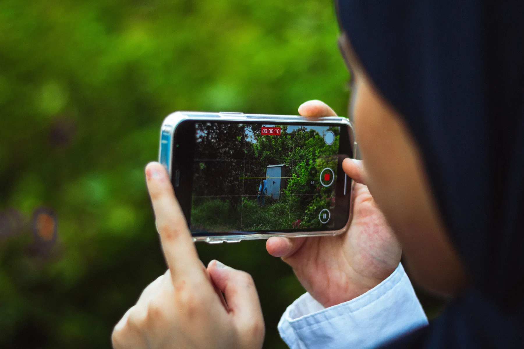 A person's hands holding up a smartphone recording a 10-second video of a garden shed and greenery, shown through the phone's camera viewfinder with a red recording indicator