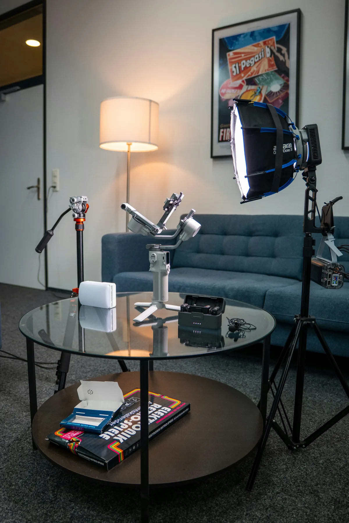 A living room set up for video production, with a camera gimbal, a softbox LED light on a stand, and a small tripod arranged on a glass coffee table in front of a couch