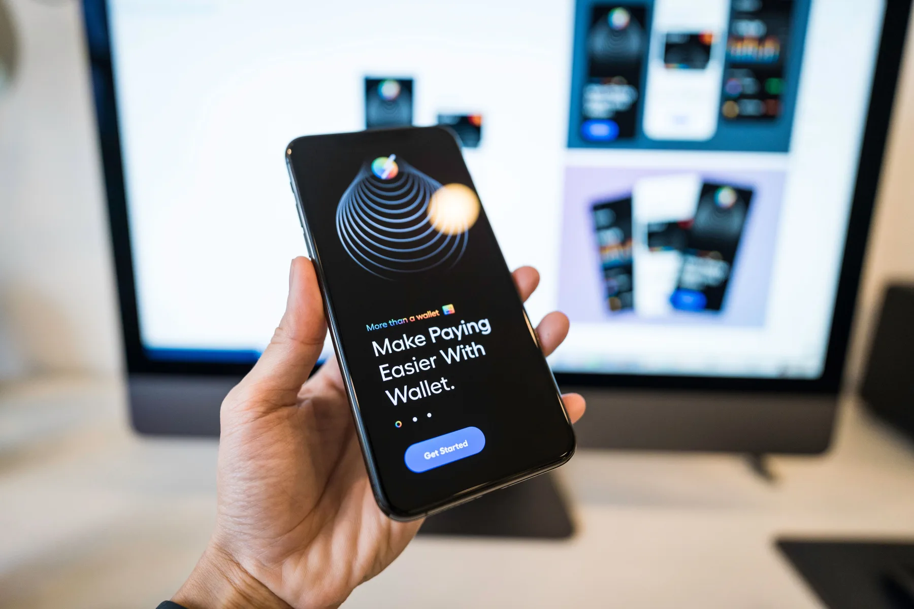 A hand holding a smartphone displaying a payment app onboarding screen that reads 'Make Paying Easier With Wallet,' with app mockups visible on a monitor in the background