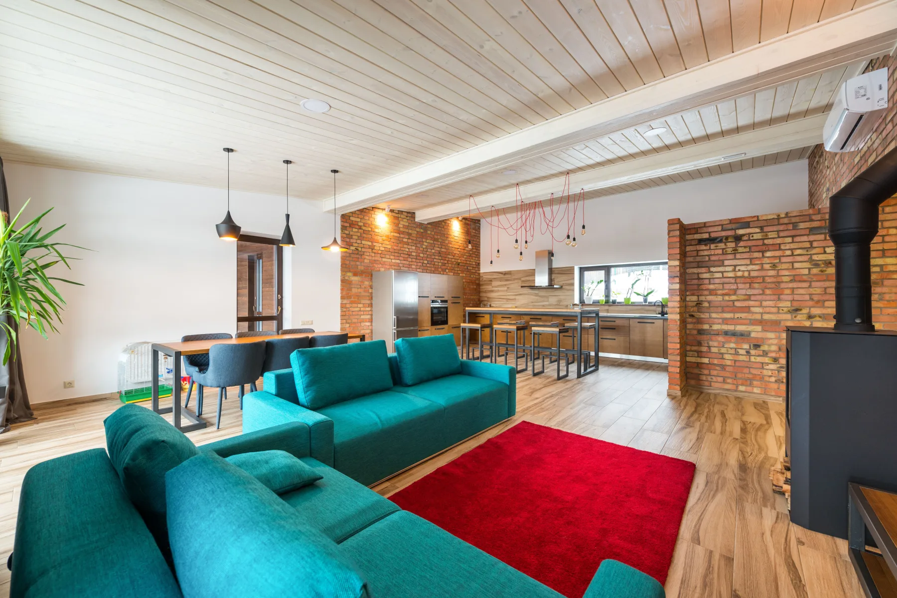 Open-plan living room with teal sofas, a red area rug, exposed brick walls, and a wood-paneled kitchen in the background