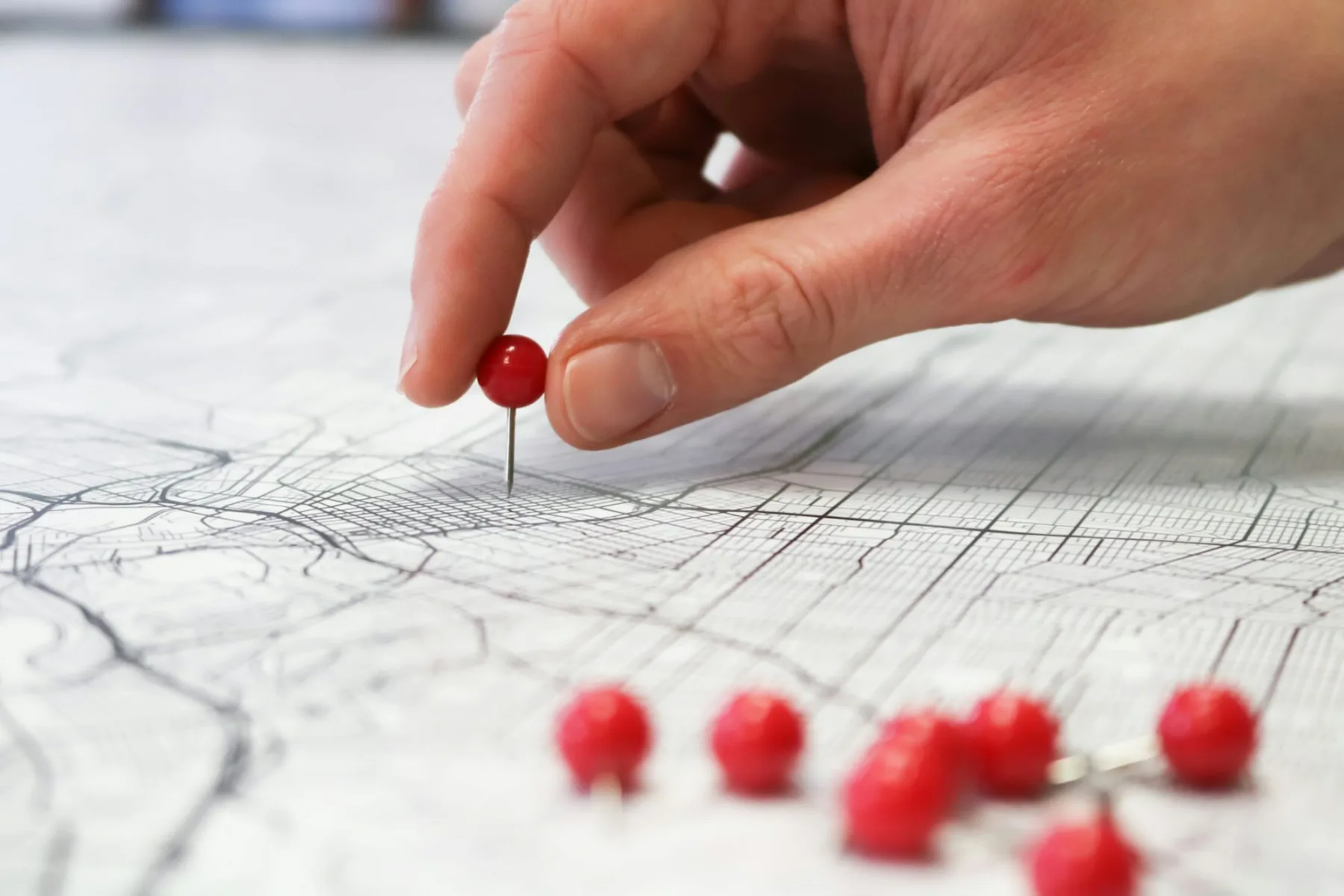 A hand pressing a red pushpin into a printed street map, with several more red pins scattered nearby