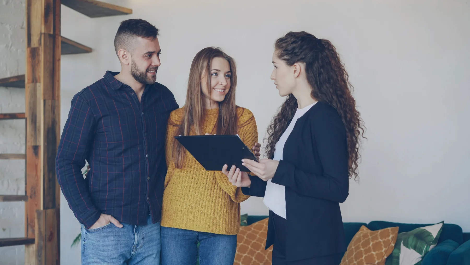 A couple standing indoors discussing a property with an agent, who is holding a dark tablet