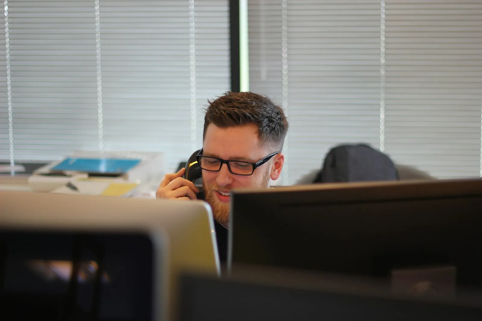 Man wearing glasses talking on an office phone at his desk, with two computer monitors in front of him