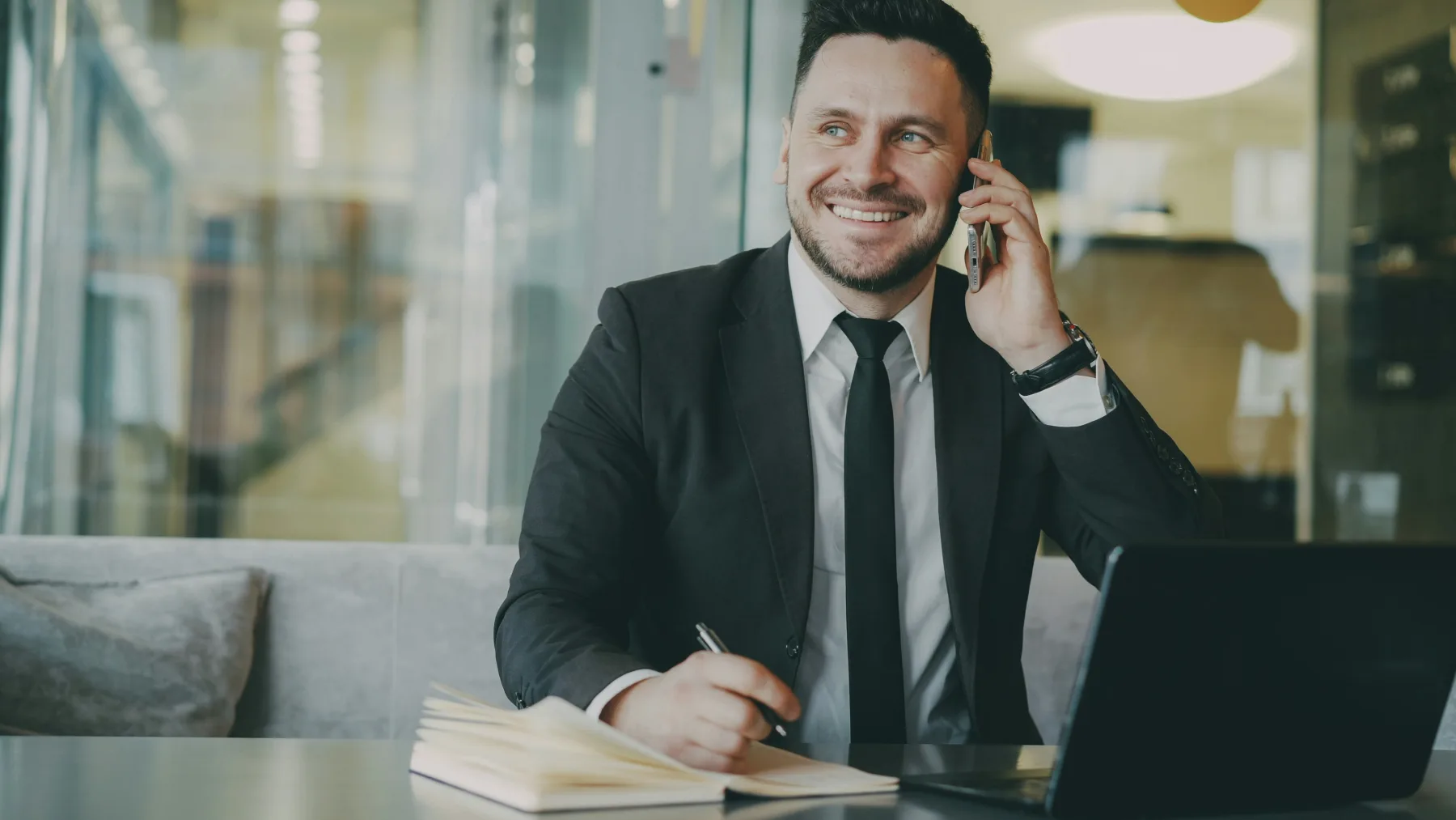 Man in a suit smiling while talking on his phone and taking notes in a notebook at his desk