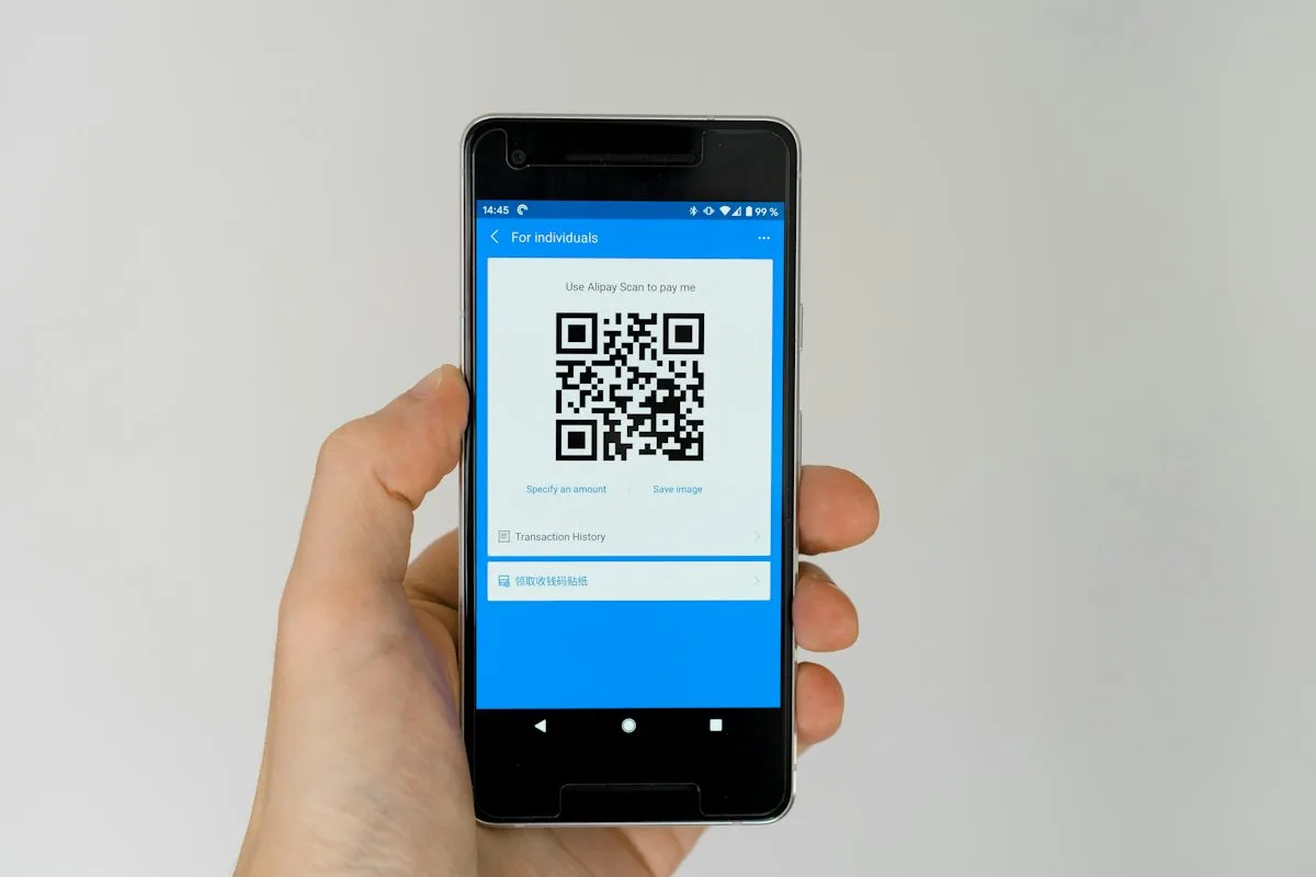 A hand holding a smartphone with a plain grey background, the screen showing a QR code on a blue app interface