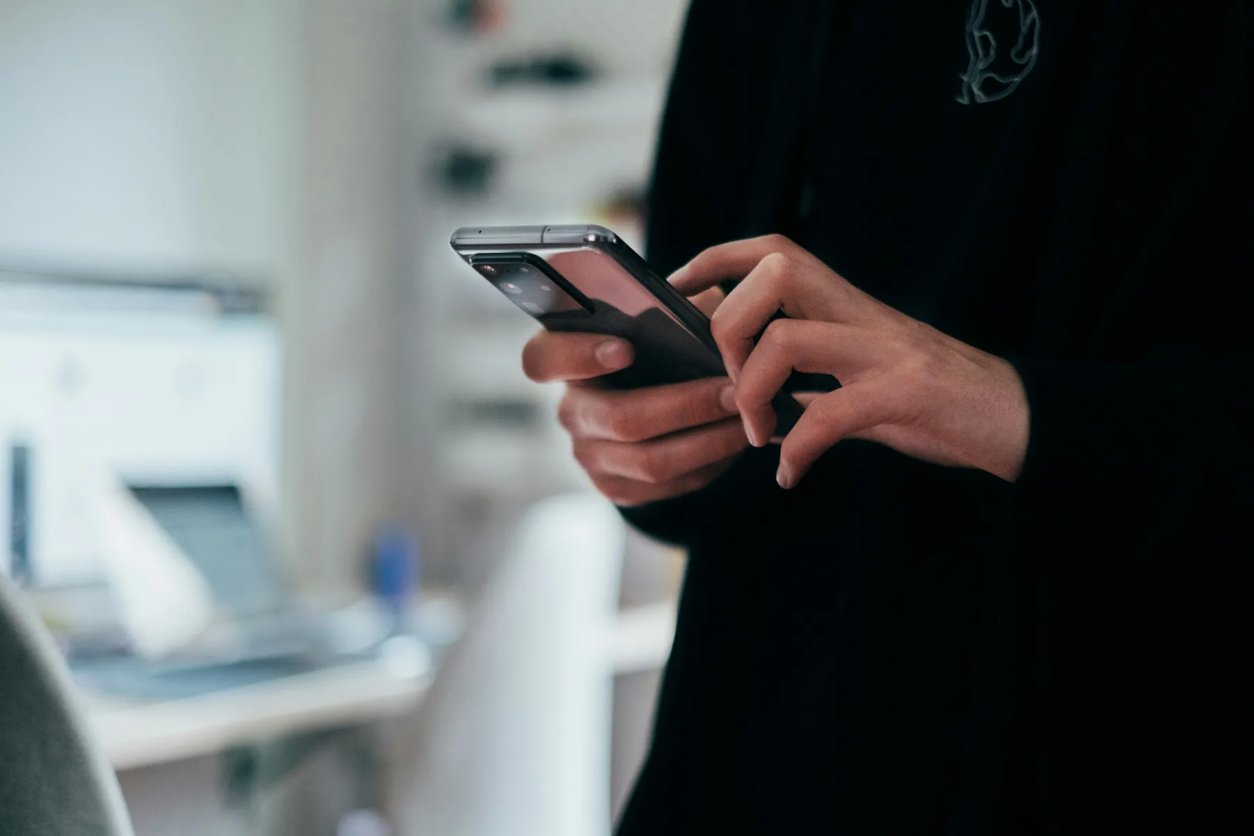 Close-up of a person's hands holding a smartphone and tapping the screen, with a blurred home office desk and monitor in the background