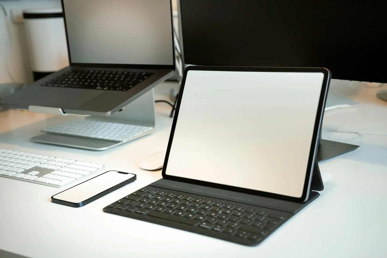 A tablet on a stand next to a laptop, keyboard, mouse, and smartphone arranged on a white desk, all with blank screens