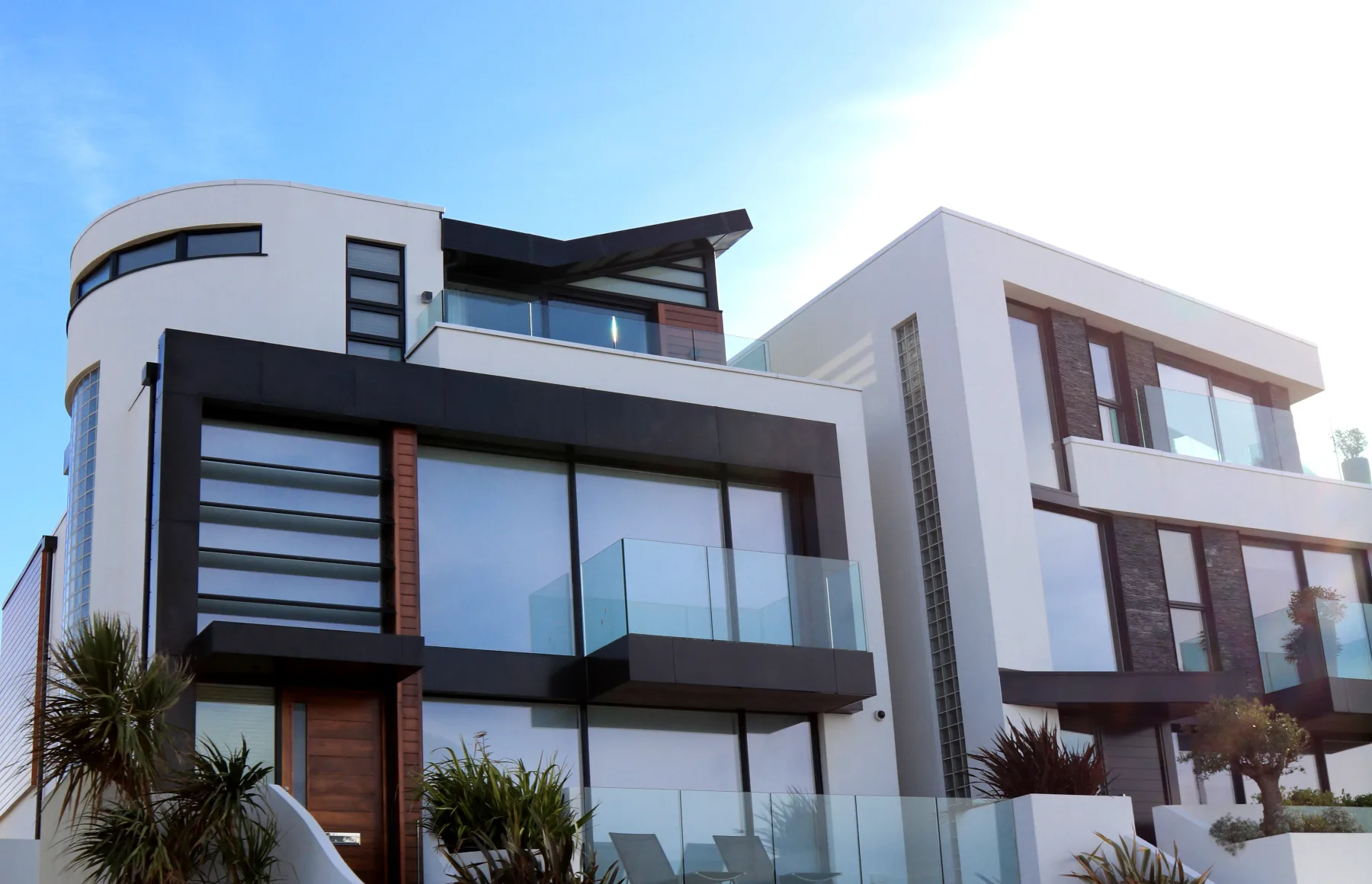 A modern multi-story house with a curved white facade, floor-to-ceiling glass windows, and glass-railed balconies under a blue sky
