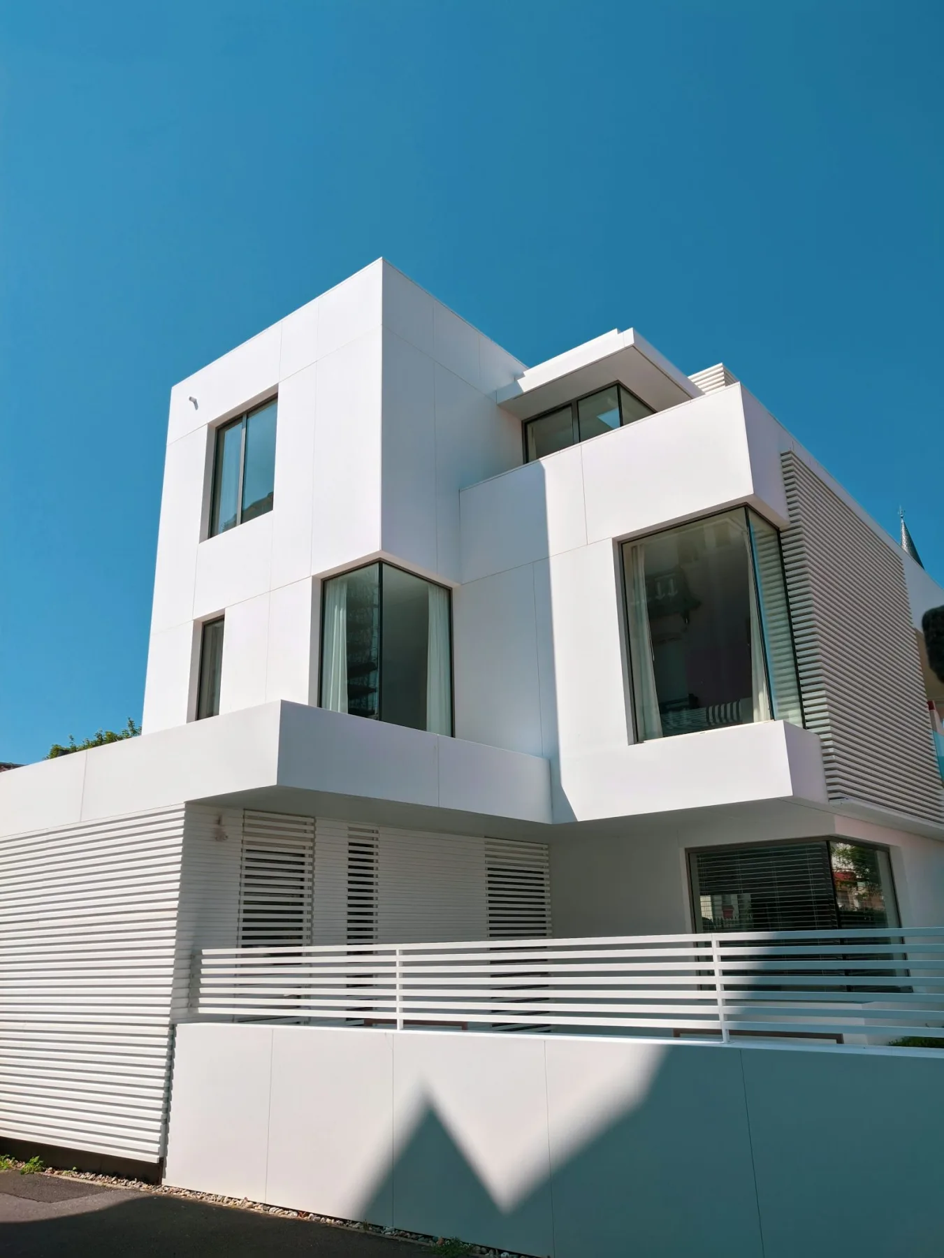A crisp white modern house with sharp angular lines, large glass windows, and a slatted privacy fence under a clear blue sky